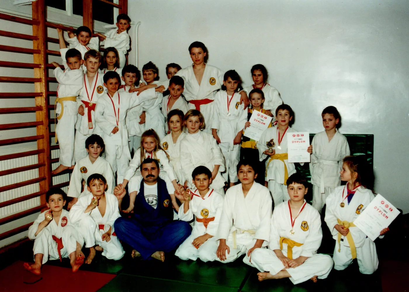 Zdjęcie grupowe dzieci w białych strojach judo z kolorowymi pasami, trzymających dyplomy i medale, z trenerem w niebieskim stroju na pierwszym planie.
