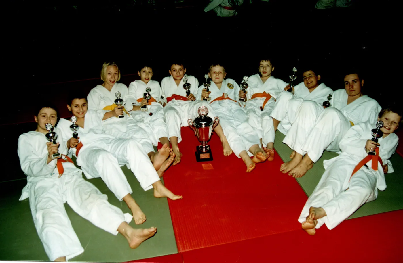 Zdjęcie dzieci w białych strojach judo siedzących na macie i trzymających puchary.