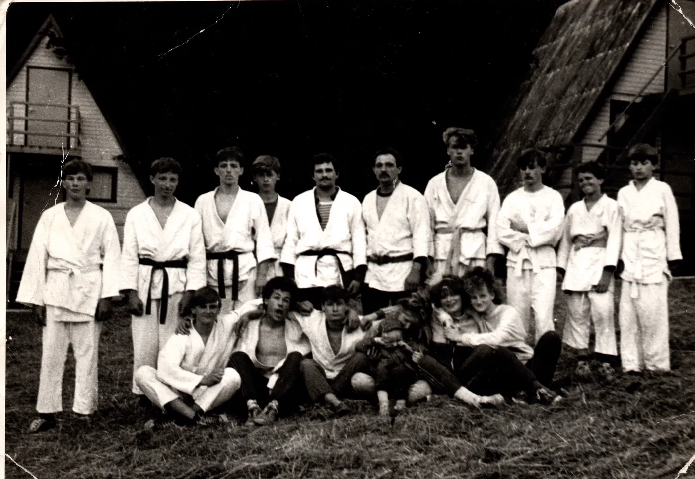 Czarno-białe zdjęcie grupy zawodników w białych strojach judo na trawie przed budynkiem z dachem kocowym.