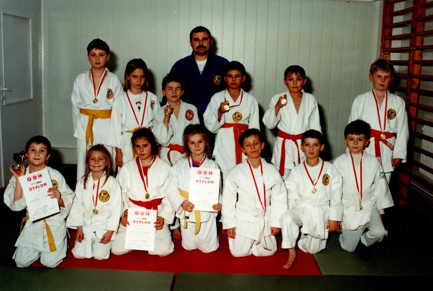 Zdjęcie grupowe dzieci w białych strojach judo z pasami, trzymających dyplomy i medale, z trenerem stojącym za nimi.